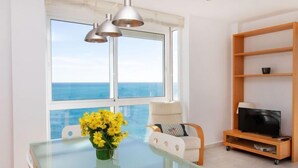 A modern living room with a glass table, a vase of yellow flowers, a TV on a wooden stand, and a view of the sea through large windows. - Apartment Gala I by Interhome (Miami Platja)