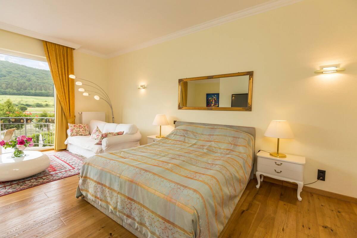 A bedroom with a large bed, bedside tables, a mirror, and a view of a balcony with flowers.