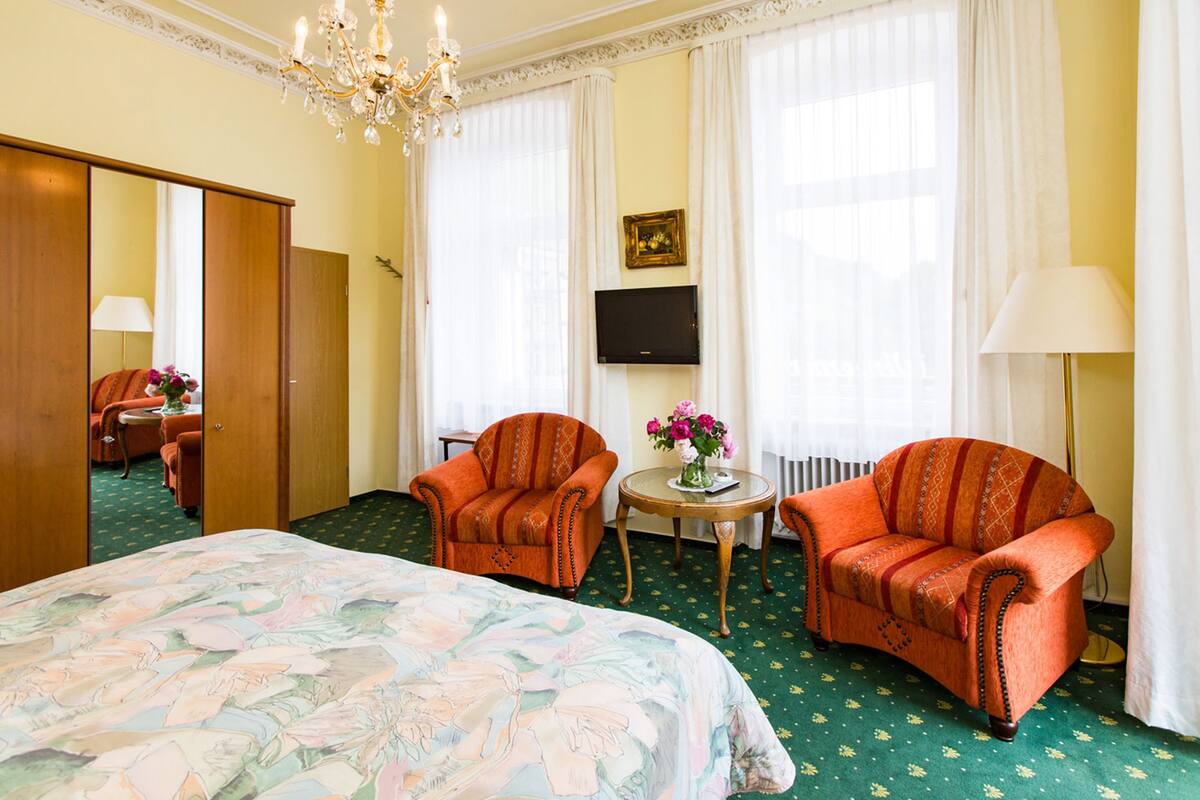 A hotel room with a large bed, two armchairs, a small table with flowers, a TV on the wall, and a chandelier.