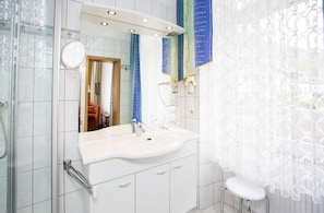 A bathroom with a white sink, mirror, and toilet. There is a shower area with a glass door and a towel rack. - Hotel Winzerverein (Zeltingen-Rachtig)