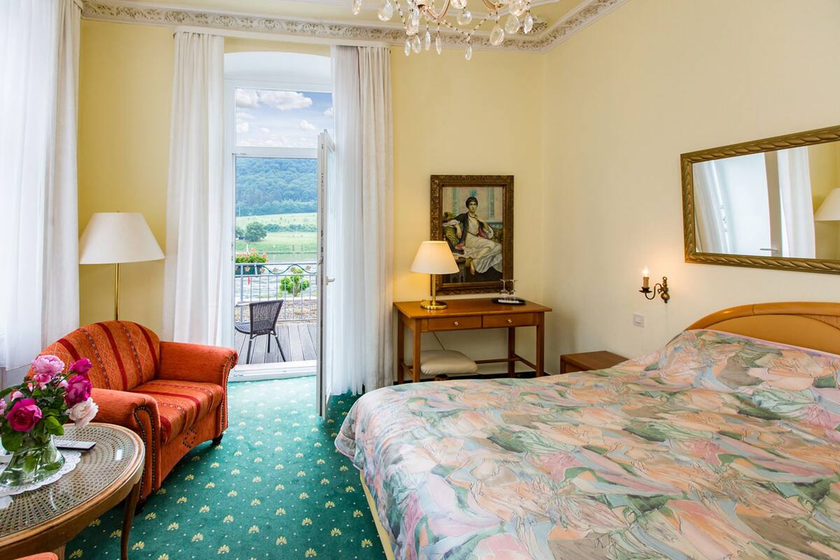 A hotel room with a bed, a chair, a table with flowers, a desk with a lamp, a painting, and a balcony with a view.