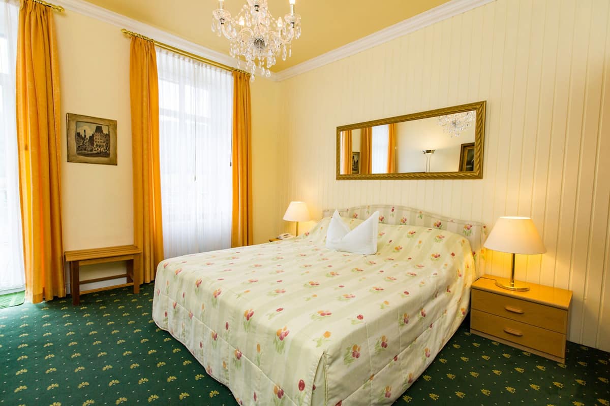 A hotel room with a bed, two bedside tables, a nightstand, a chandelier, a mirror, and a window with curtains.