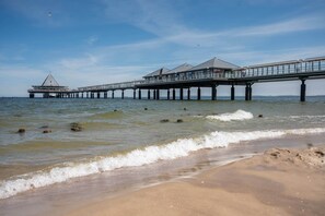 A pier extending into the water with buildings on it, a sandy beach in the foreground, and a clear sky.