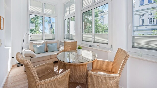 A bright room with a glass-top table surrounded by wicker chairs, a sofa with cushions, and large windows with blinds. - Cozy vacation apartment in Heringsdorf close to the beach (Seebad Heringsdorf)