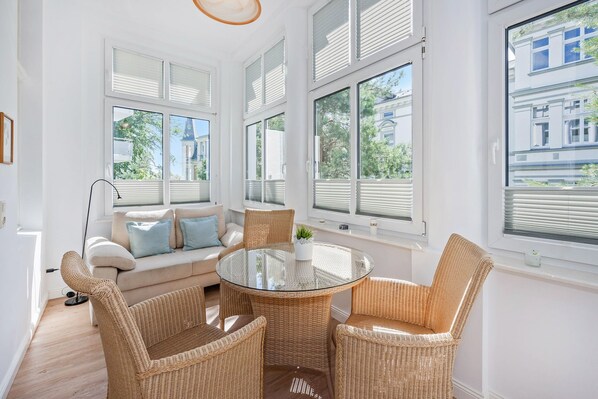 A bright room with a glass-top table surrounded by wicker chairs, a sofa with cushions, and large windows with blinds.