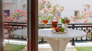 A round table with two glasses of amber liquid, a glass of red liquid, and a plate with an apple and a green fruit, set against a balcony with pink flowering plants and a white building in the background. - Naxos Diamond Studios Apartments (Naxos)