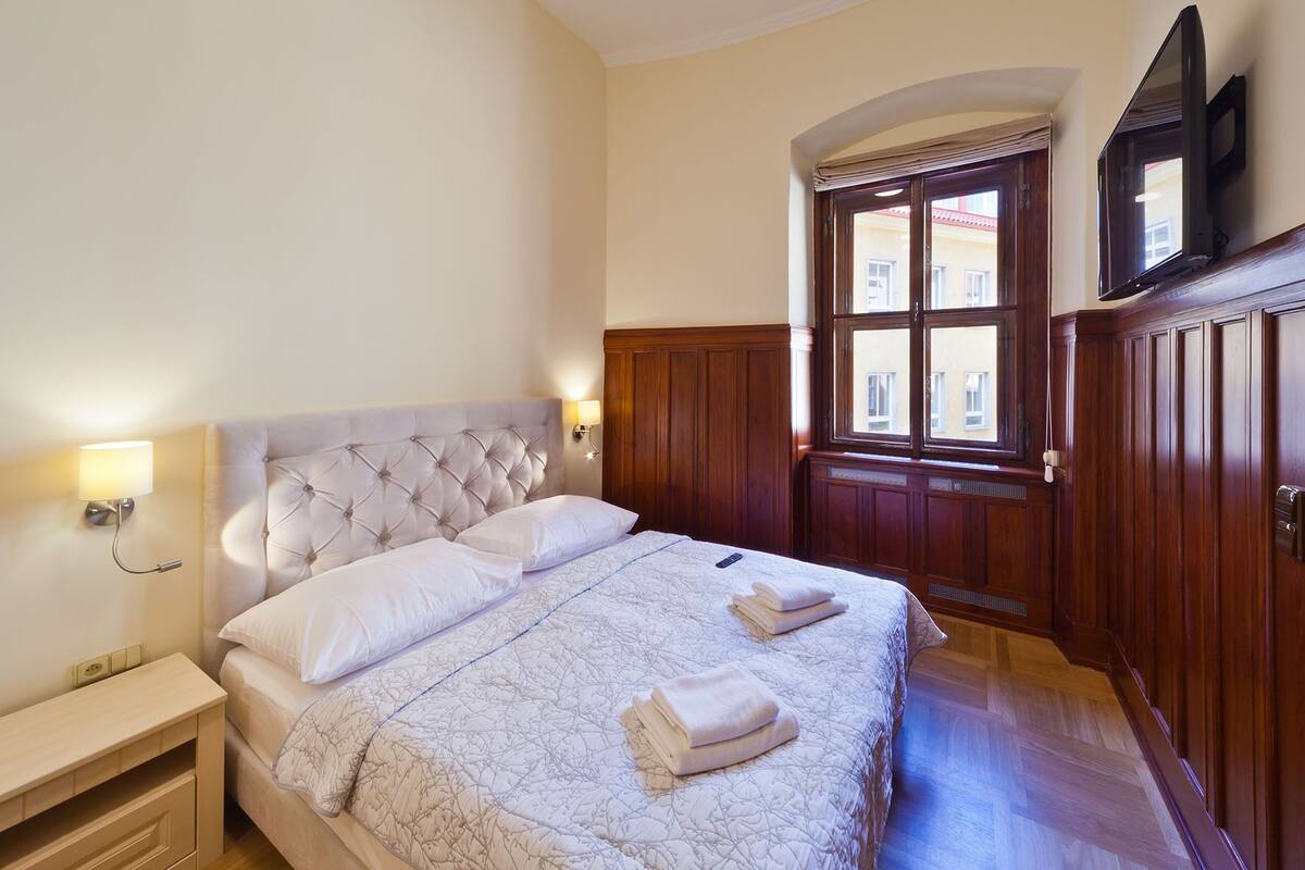 A neatly arranged bedroom with a large bed, a bedside table, a TV mounted above the door, and a view of the street outside.