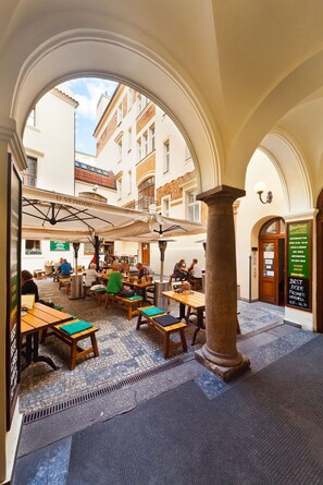 An indoor café with wooden tables and chairs, a stone column, and a view of buildings through an archway. - Residence u Vejvodů (Prague)