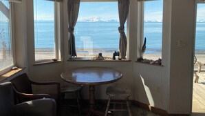 A room with a round wooden table and two chairs, a view of a beach with mountains in the distance, and a window with curtains. - Perfectly Positioned to Play on the Kenai Peninsula (Ninilchik)