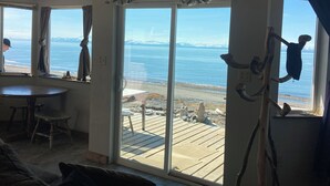 A room with a large sliding glass door leading to a deck with a view of a beach and mountains. There is a table and chairs inside the room. - Perfectly Positioned to Play on the Kenai Peninsula (Ninilchik)