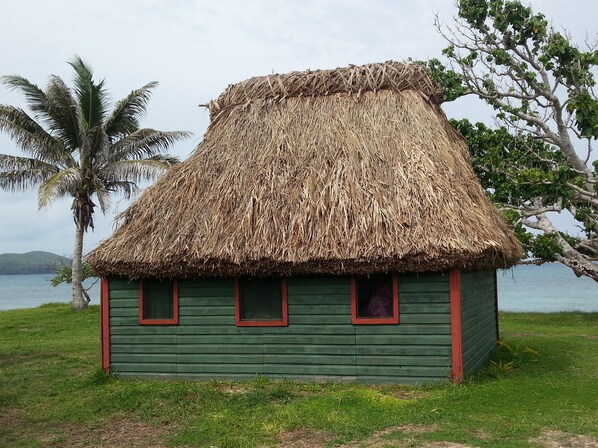 Una cabaña con techo de paja, fachada verde y marcos de ventana rojos, ubicada en un área cubierta de pasto cerca de agua y palmeras.