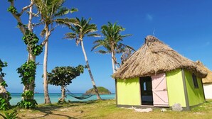 A small thatched-roof hut with a pink door, surrounded by palm trees and a hammock, overlooking a clear blue sea. - Traditional Bure by the beach in the authentic Yasawa Village (Yasawa islands)
