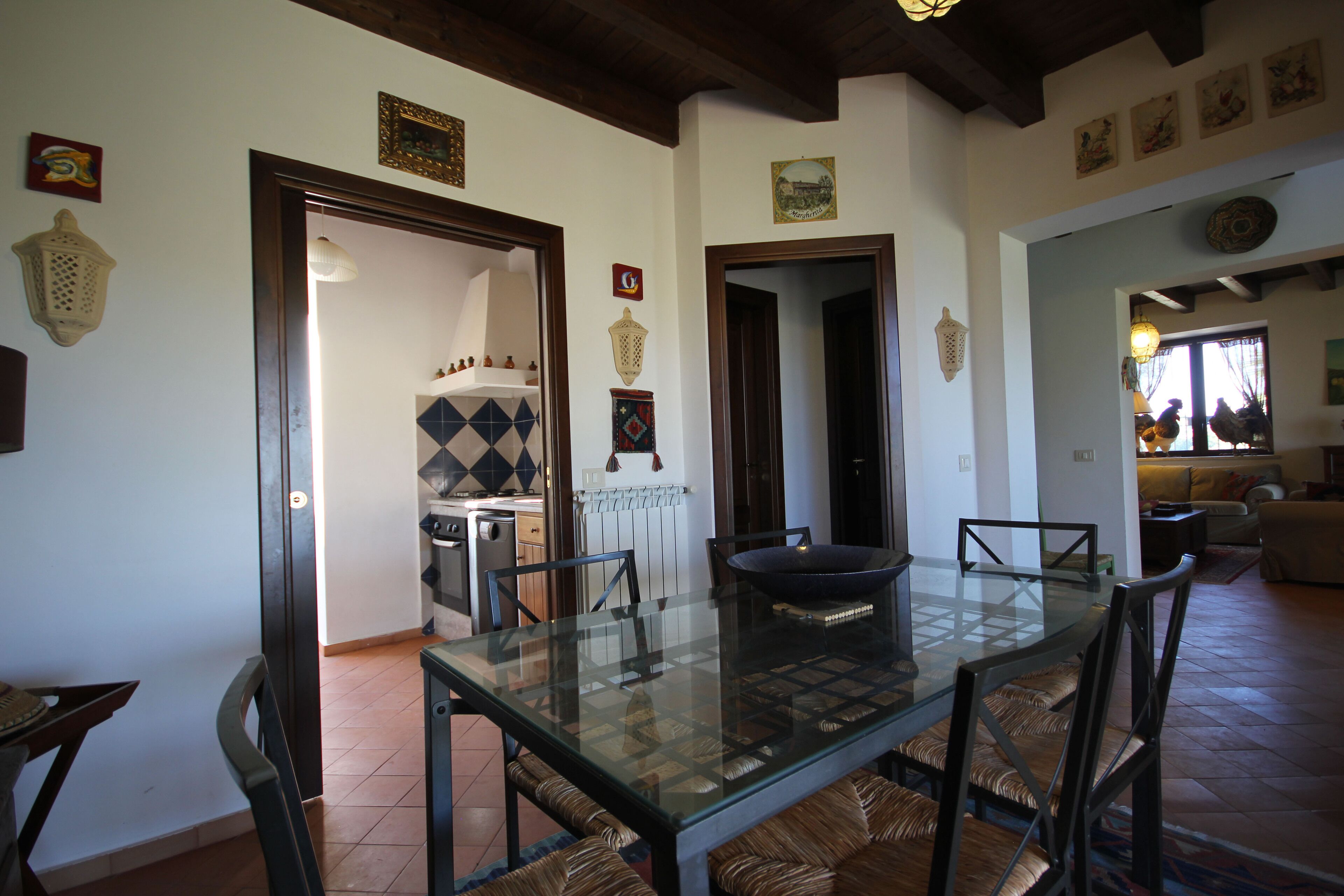 A dining area with a glass table and wicker chairs, a kitchen in the background, and a living room with a sofa and a window.