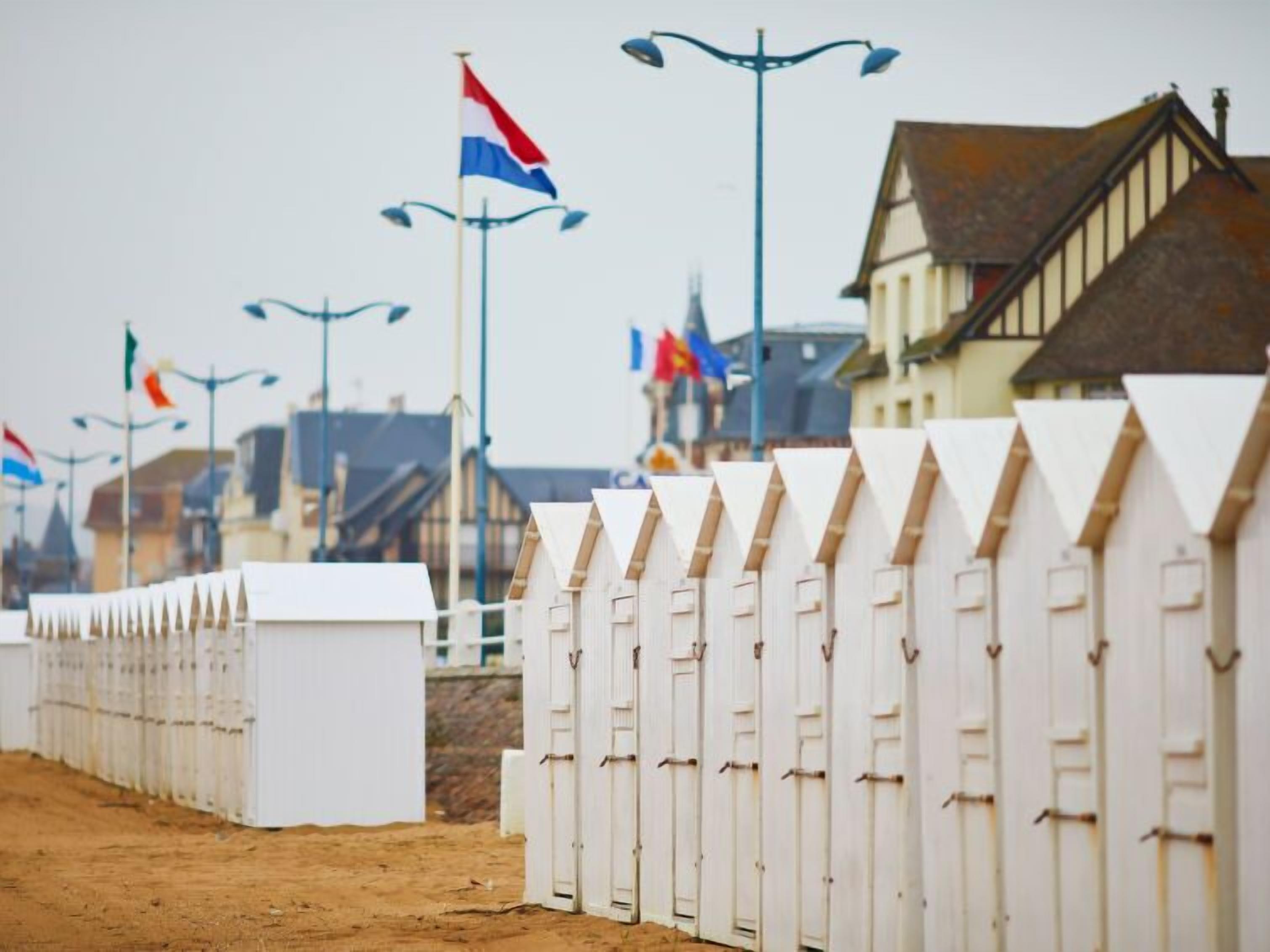 A row of white beach huts with a sandy path leading to them, set against a backdrop of buildings and flags.