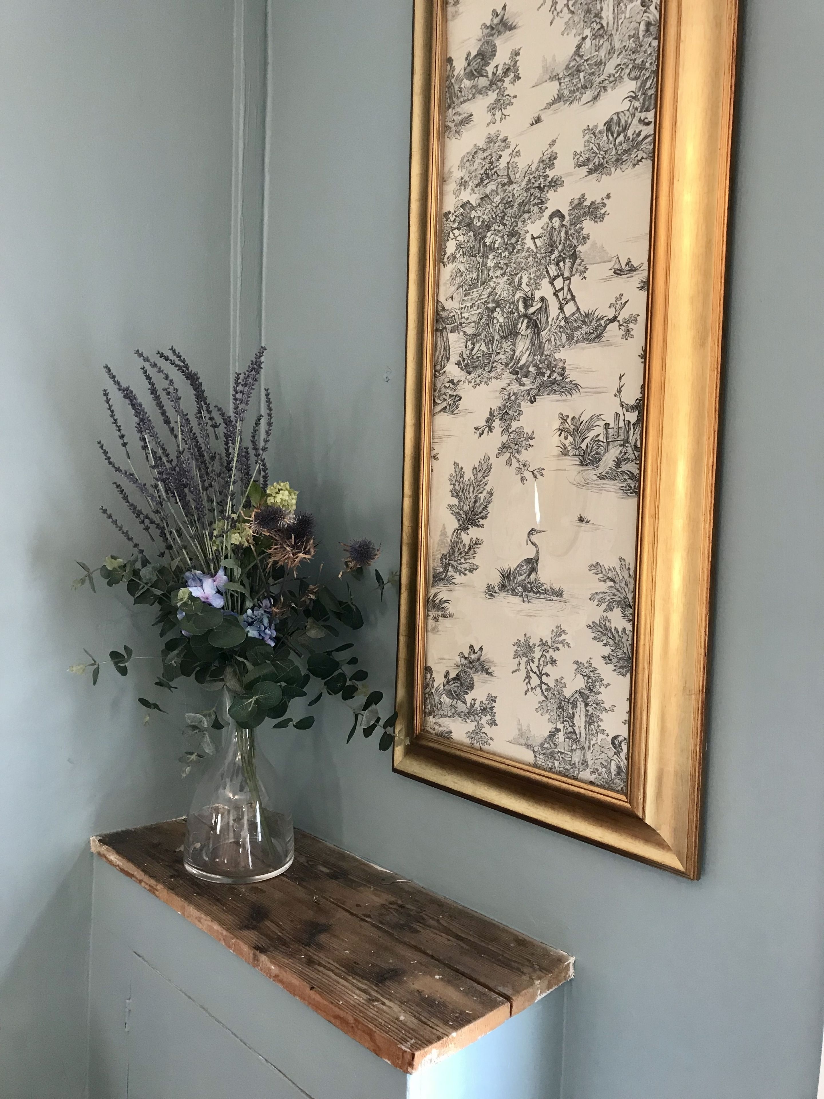 Una ilustración botánica en un marco en la pared, un florero con flores en un estante de madera y una pared azul claro.