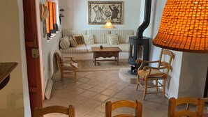 Uma sala de estar acolhedora com sofá, mesa de centro e mesa de jantar com cesta de frutas. Há uma lareira e um abajur no aposento.
