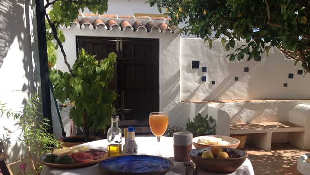Uma área de refeições ao ar livre com uma mesa posta para o café da manhã, incluindo pratos de frutas, pão e um copo de suco de laranja.