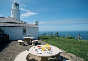 Mesa de jantar posta com pratos, copos e talheres, com vista para a costa e um farol ao fundo.