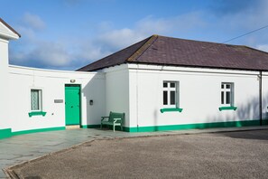Um edifício branco com porta e janelas verdes, um banco na frente e um espaço pavimentado.