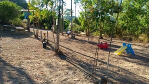 A playground with swings and a slide, surrounded by trees and a dirt ground.