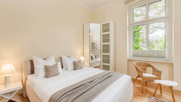Un dormitorio con cama, mesitas de noche, una silla, ventana y espejo.