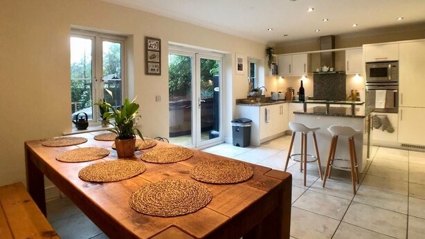 A modern kitchen with a wooden dining table, bar stools, and a large window overlooking a garden.