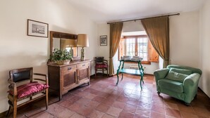 Ein Zimmer mit einem hölzernen Schminktisch, einem grünen Sessel, einem roten Stuhl, einem Tisch mit Lampe und einem Fenster mit Vorhängen.