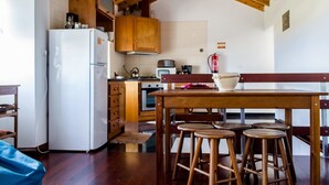 A compact kitchen with wooden cabinets, a white refrigerator, a wooden dining table with stools, and a wooden ceiling. - Casa da Avó - Quinta da Ribeira da Urze (Prainha de cima)