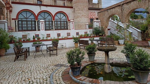 Un patio con suelo de adoquines, una fuente, plantas en macetas y un edificio con ventanas de arco y una torre.