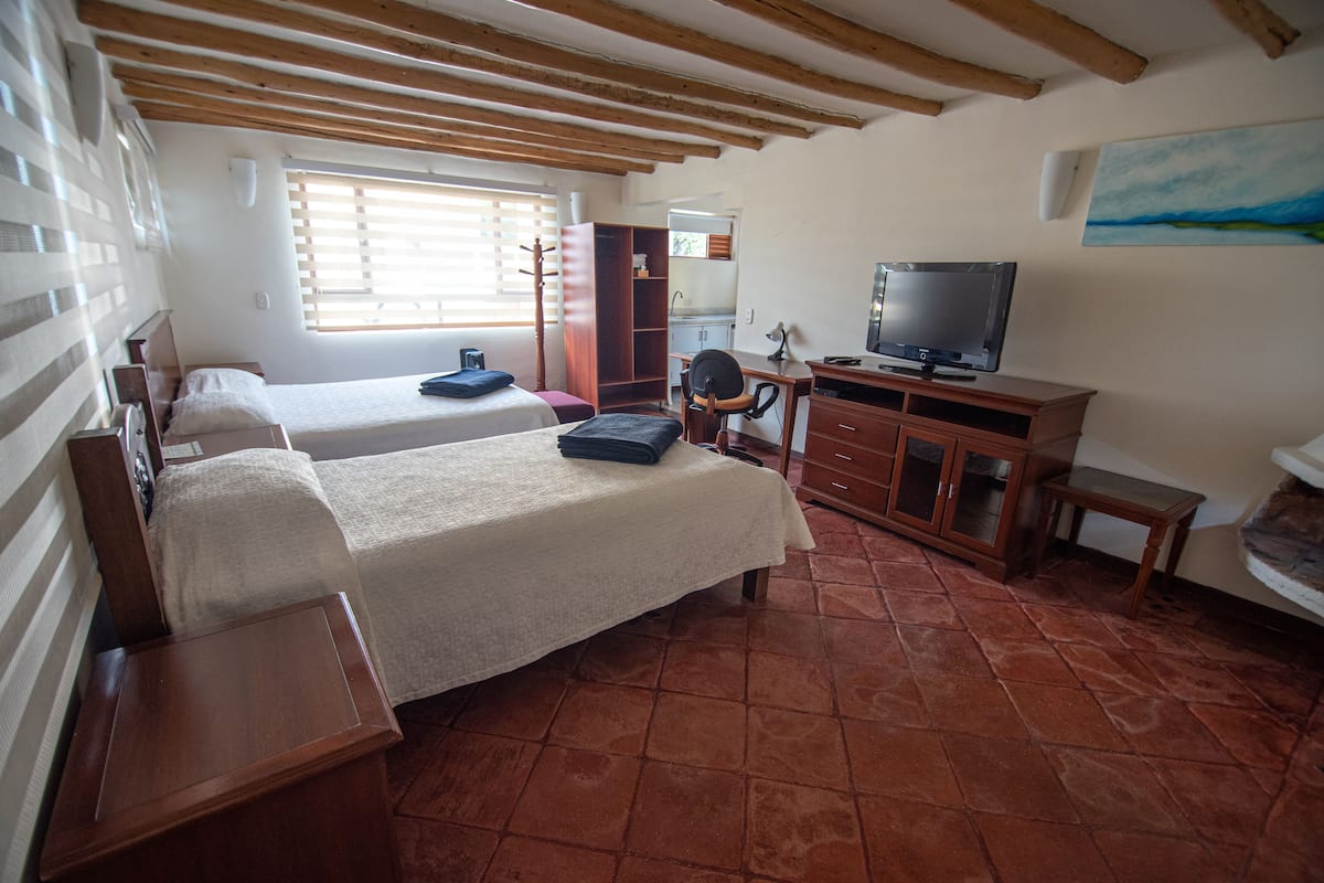 A room with two beds, a desk with a television, a chair, and a window with blinds.
