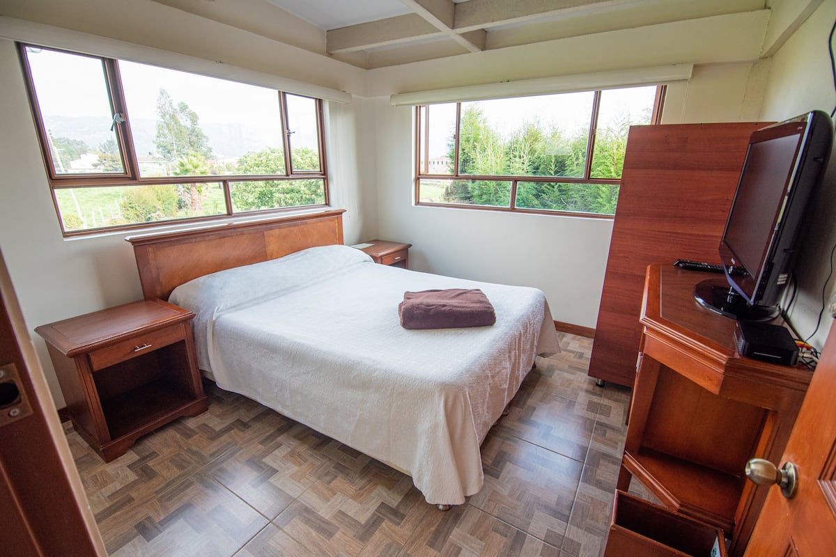 A bedroom with a bed, wooden nightstands, a TV, and large windows.