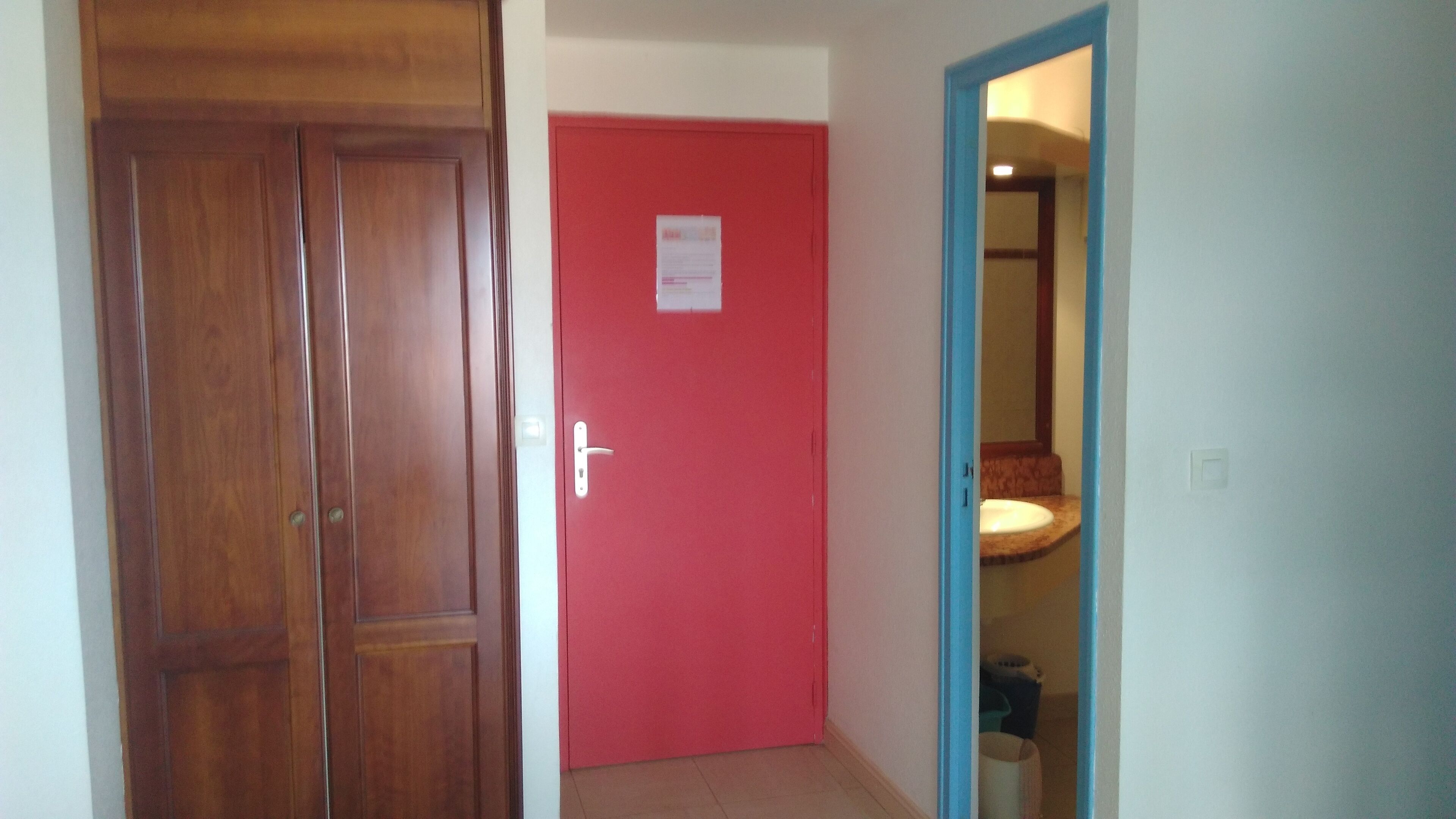 Um corredor com uma porta de madeira, uma porta vermelha e um banheiro visível através de uma porta aberta.