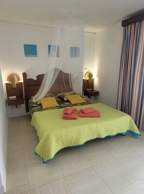 A bed with a canopy, two bedside tables with lamps, a striped curtain, and a tiled floor. - Studio Vue mer des Caraïbes, un Petit Coin de Paradis (Sainte Anne)