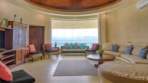 Amplia sala de estar con techo curvo, un ventanal con vista al océano y diversas opciones de asientos, incluyendo sofás y sillones.