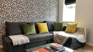 Een moderne woonkamer met een grijze bank, groene en gele kussens, een glazen salontafel met boeken en behang met een patroon.