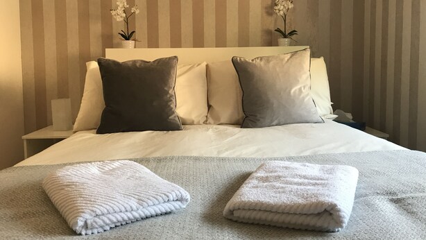 Een keurig opgemaakt bed met twee kussens, een nachtkastje en twee potorkideeën op het hoofdeinde.