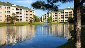 A multi-story building with balconies, a pond with a fountain, and a tree in the foreground.