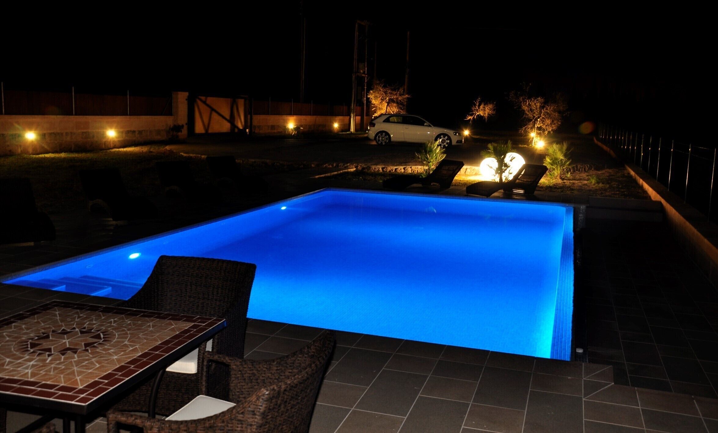 Een zwembad met verlicht blauw water, omringd door tuinmeubels en een nabijgelegen auto.