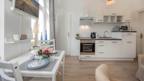 Uma cozinha moderna com armários brancos, uma sala de jantar com mesa posta para dois e uma sala de estar com sofá.