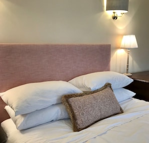 A neatly made bed with white linens and a decorative throw pillow, a wall-mounted light fixture, and a bedside table with a lamp.