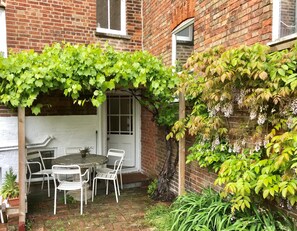 A brick building with a white door and window, a round table with chairs, and a garden area with green foliage and white flowers.