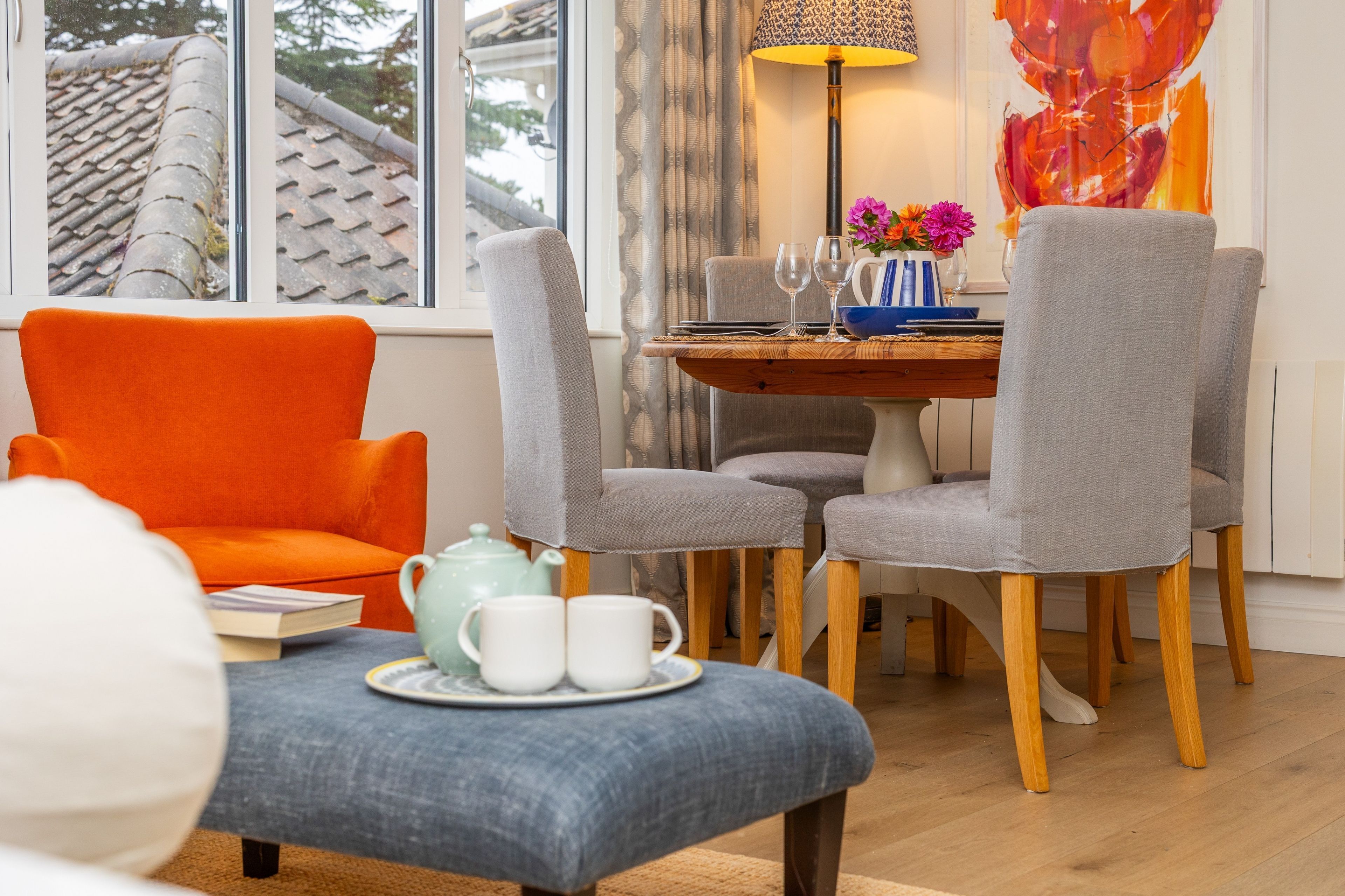 A room with a dining table, chairs, a sofa, a lamp, and a window with a view of a tiled roof.