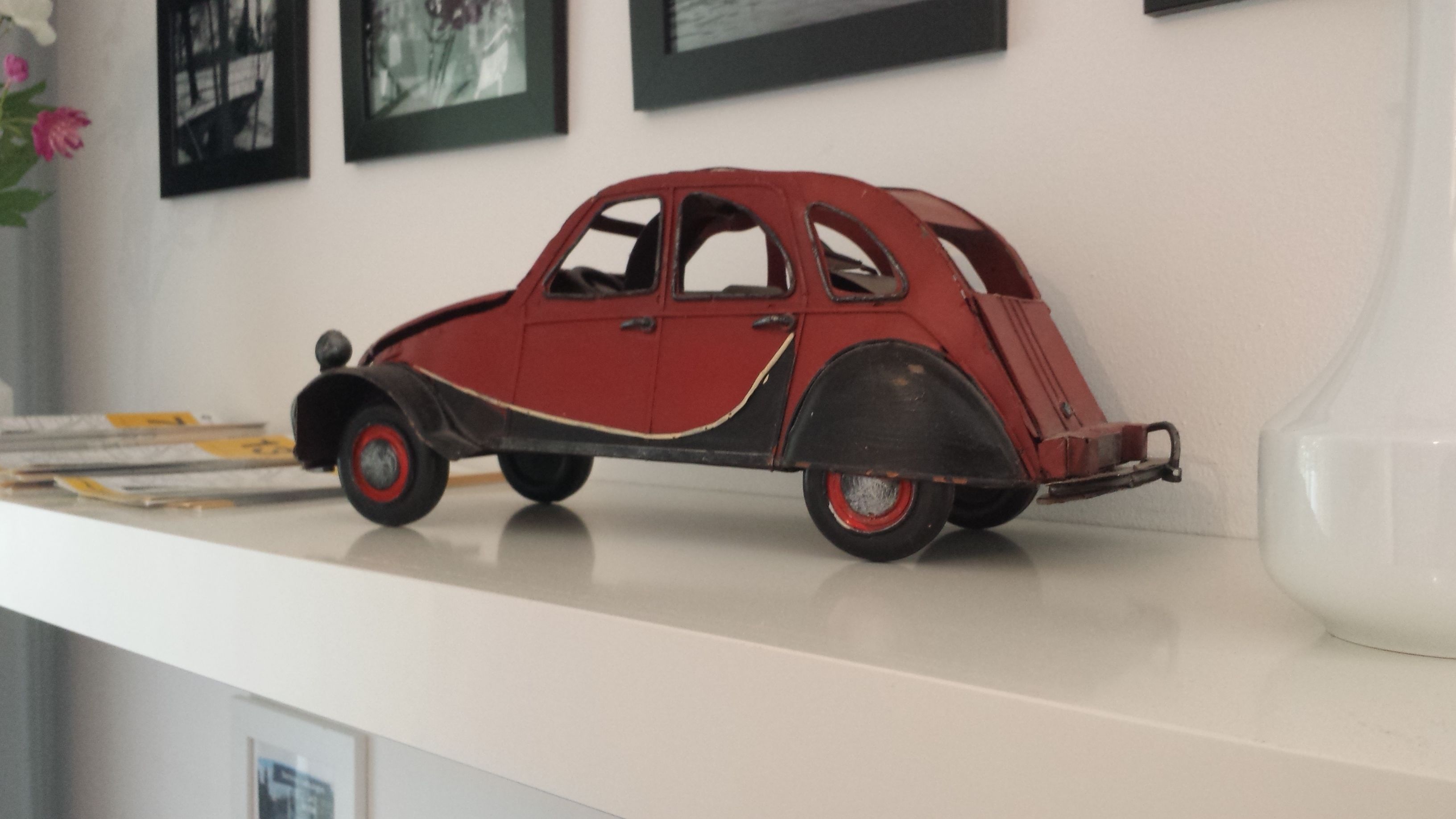 Ein altes Automodell auf weißem Untergrund, mit einer weißen Vase und gerahmten Bildern im Hintergrund.