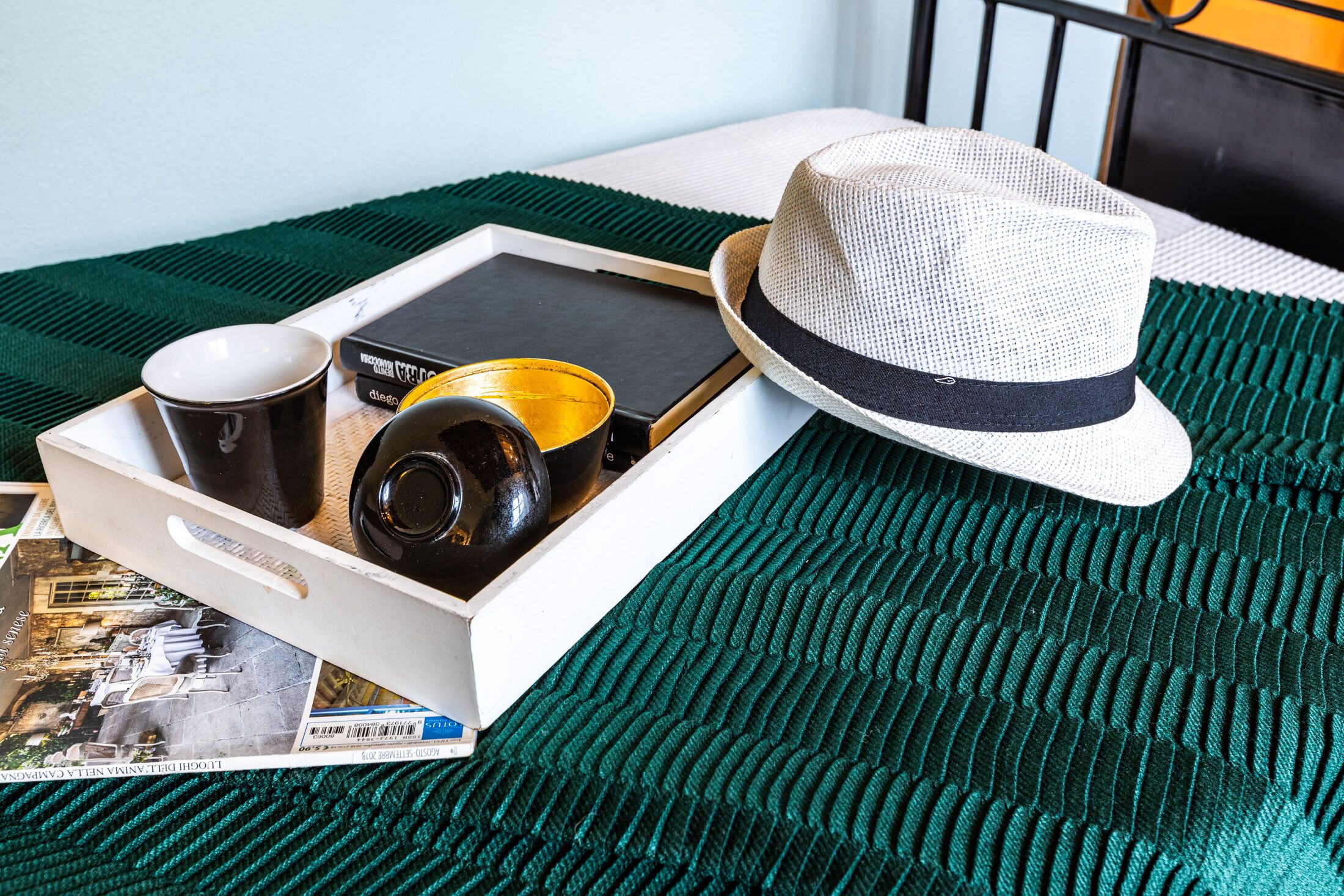 Una bandeja con un libro, dos tazas, una vela y un sombrero sobre una cama con una manta verde.