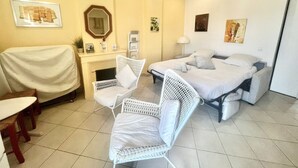 Ein Hotelzimmer mit einem Bett, zwei weißen Korbstühlen, einem kleinen Tisch, einem Schreibtisch mit Spiegel und einem Wandregal.