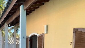 A covered outdoor seating area with wicker furniture and a glass table, adjacent to a building with a closed shutter door. - Beira Mar House with pool and waterfall in Mongaguá with 06 suites (Mongaguá)