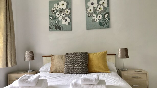 A neatly made bed with white linens and towels, a headboard with a patterned pillow, and two wall-mounted lamps. - The Woodfarm Lodge - 3 Bedroom House (Oxford)