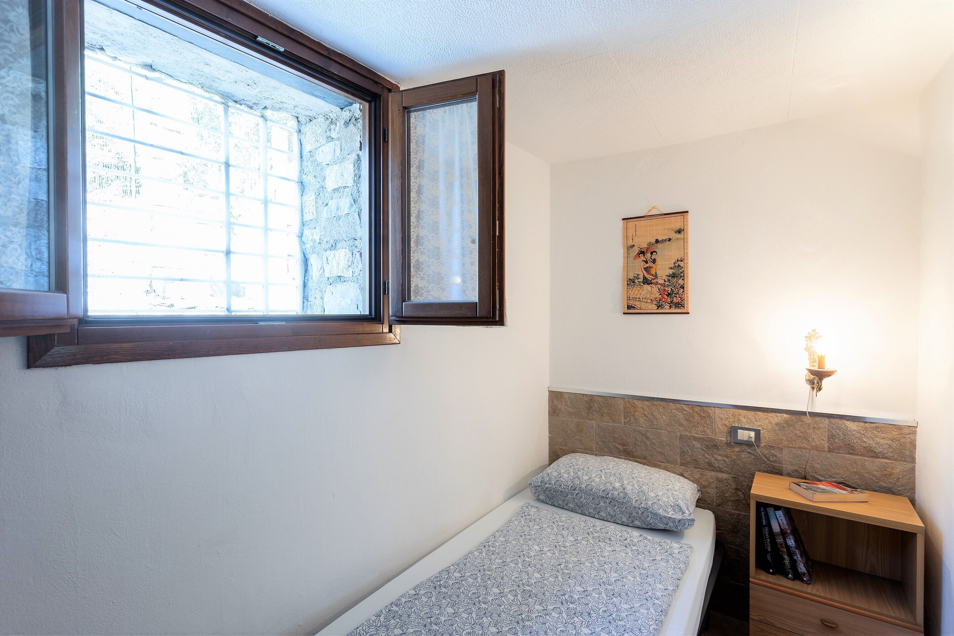 Une petite chambre avec un lit simple, une table de chevet avec une lampe, une fenêtre donnant sur un mur de pierre et un tableau accroché au mur.
