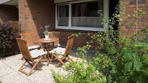 A patio with a table and four chairs, a potted plant, and a window with blinds. - Vacation apartment Strandbox (Norddeich)