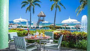 Un área de comedor junto a la piscina con sillas, una mesa, palmeras y sombrillas.